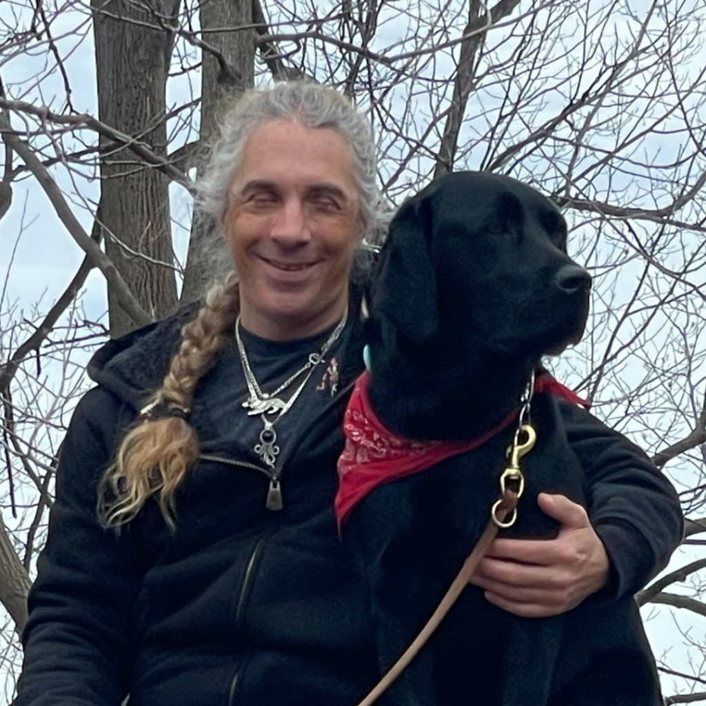 Pat McKenna smiles outside with an arm around a black Labrador with a red bandana.
