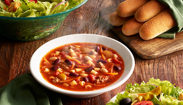 A rustic wooden table setting with a bowl of steaming Olive Garden minestrone soup, a green salad in a bowl, and a plate of breadsticks wrapped in a green napkin.
