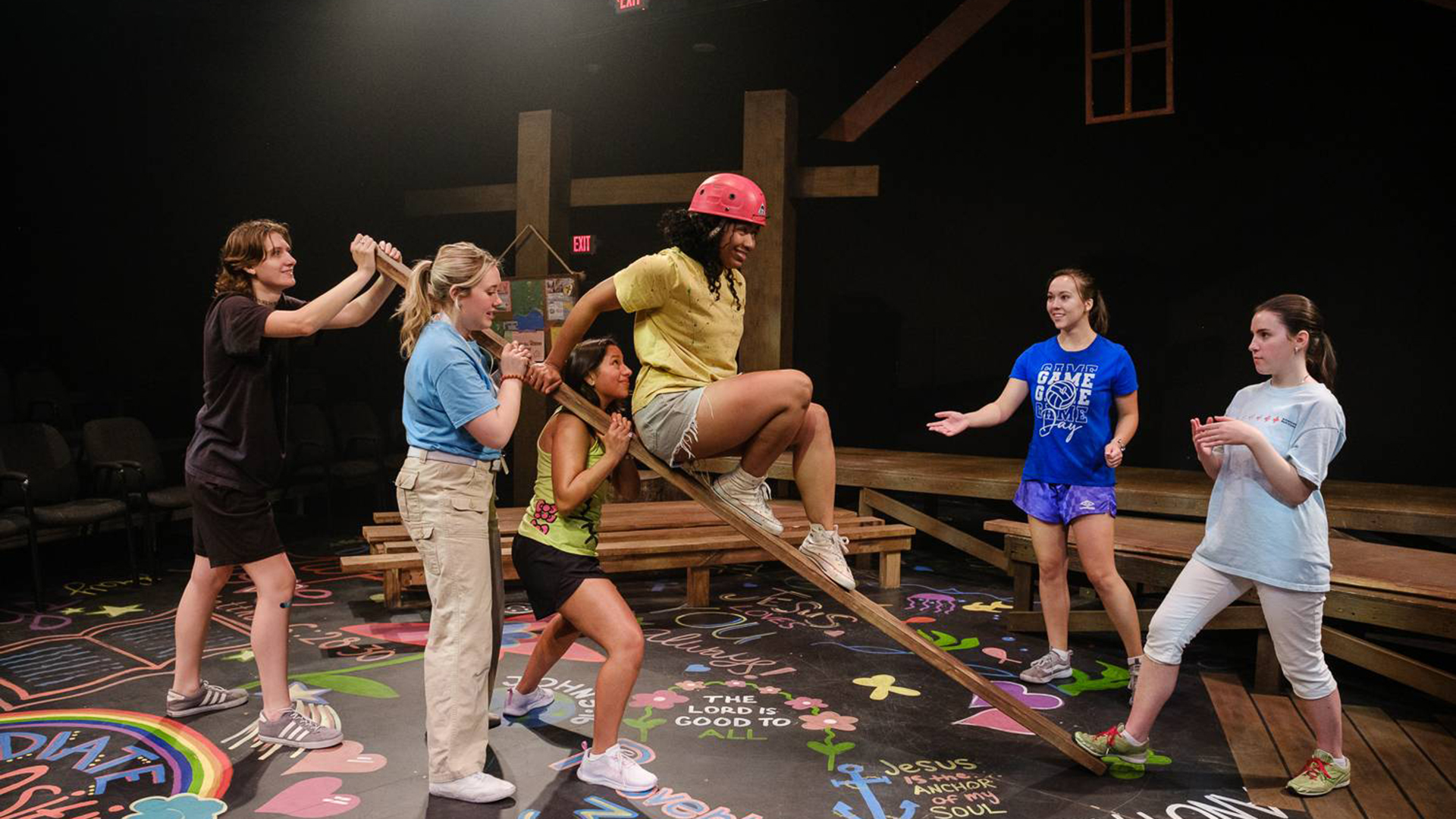 Performers in a colorful stage play scene with one person climbing a wooden beam supported by others.