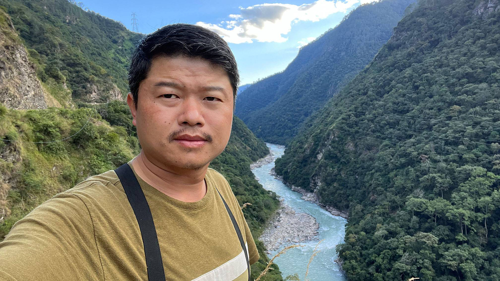 Man taking a selfie in a lush green valley, with a winding river flowing between steep, forested mountains under a partly cloudy sky.