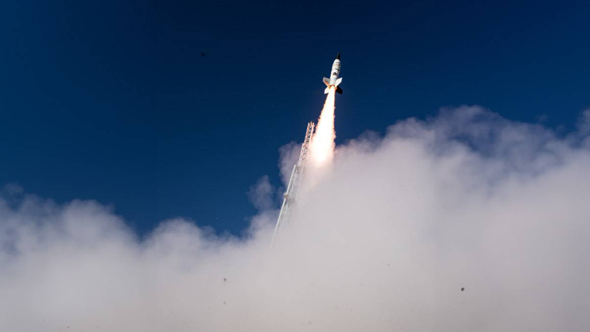 Rocket launching vertically into a clear blue sky, surrounded by a plume of white smoke at the base, with bright flames visible from its exhaust.