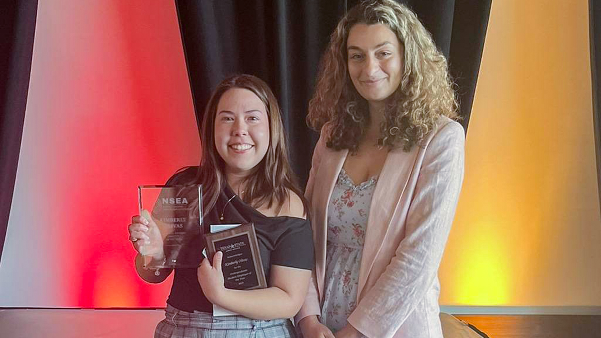 Two women standing, one is holding two Employee of the year awards