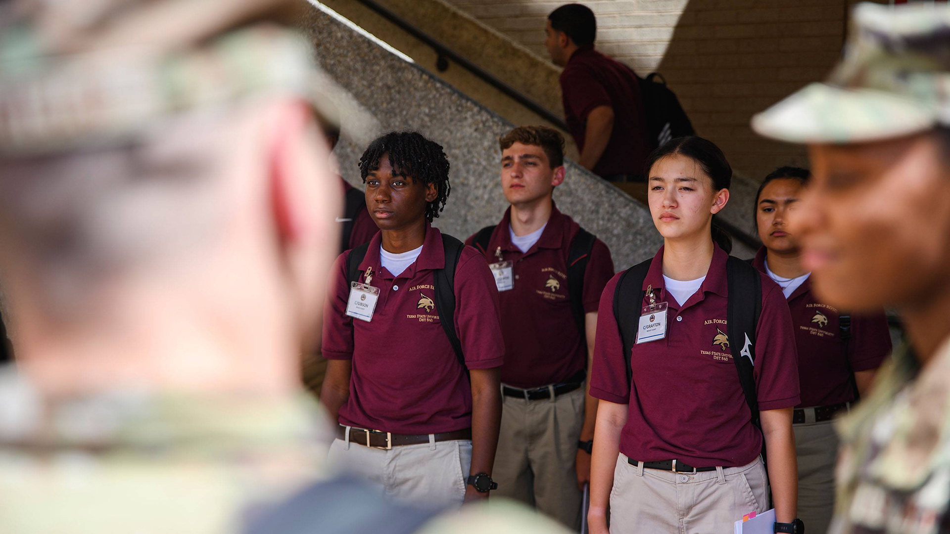 Diverse students in maroon uniforms stand at attention while being addressed by an instructor.