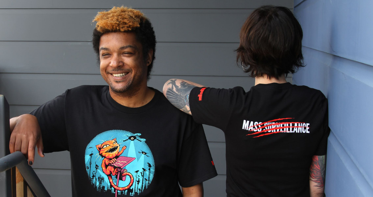 Two people wear EFF Claw Back member t-shirts. The front shows a cat swatting at spy cameras and the back says 'Mass Surveillance' with red claw marks through it.