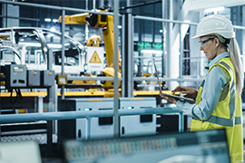 A woman wearing a yellow safety vest and a white hard hat uses a laptop computer to operate robots on an Advanced Manufacturing automobile assembly line.