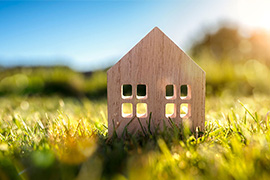 A wooden cutout of a small one-story house sits in an open field in a rural setting