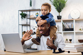 A young father attends a virtual work meeting from home with his child on his shoulders.