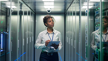 A young female worker holds a clip board and walks through a data center server room