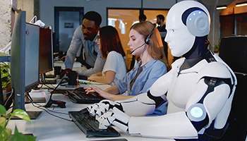 A robot types on a computer at a workstation alongside human employees