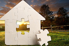 A puzzle in the shape of a house with a piece in the middle missing in front of rural farmland illustrates missing middle housing