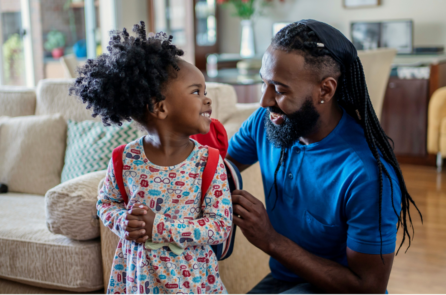 An African American male with a beard and long braids helps a young African American girl put on her backpack.