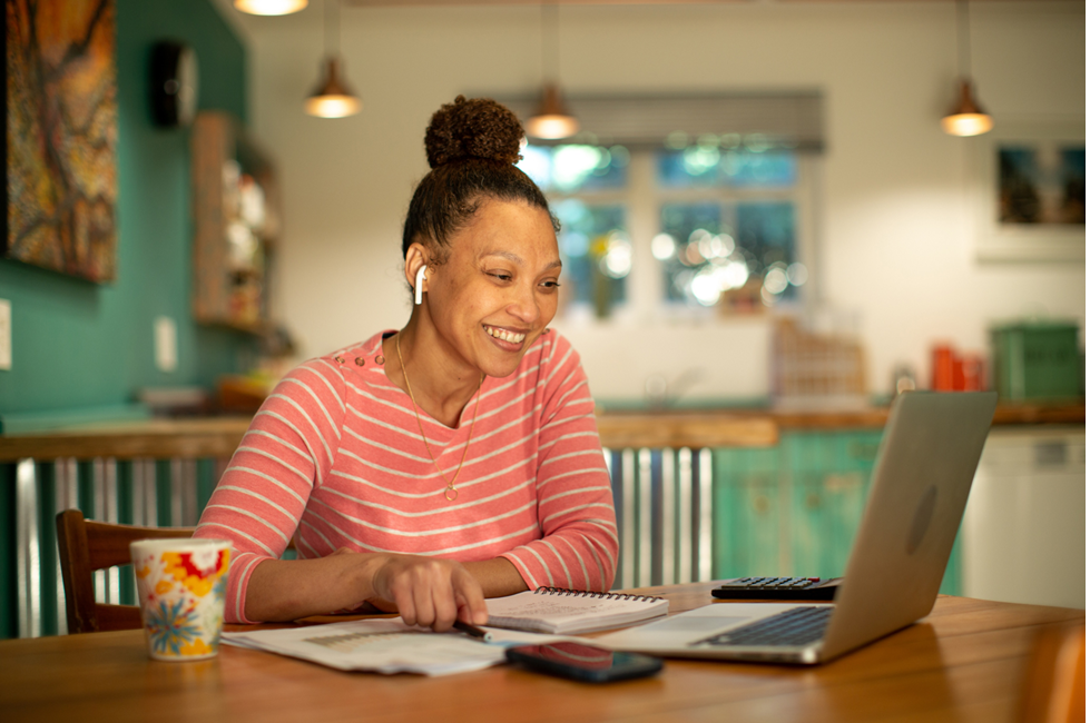 African American woman sits at a table with a notebook, cell phone, and coffee cup, looking at her laptop computer.