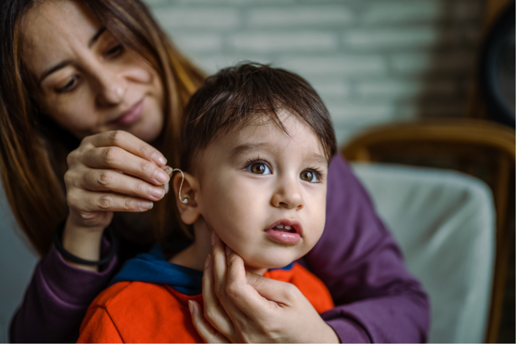 An image of an adult woman placing a hearing aid in the right ear of a young child while he is sitting on her lap. The woman is wearing a purple shirt, and the child is wearing an orange shirt.
