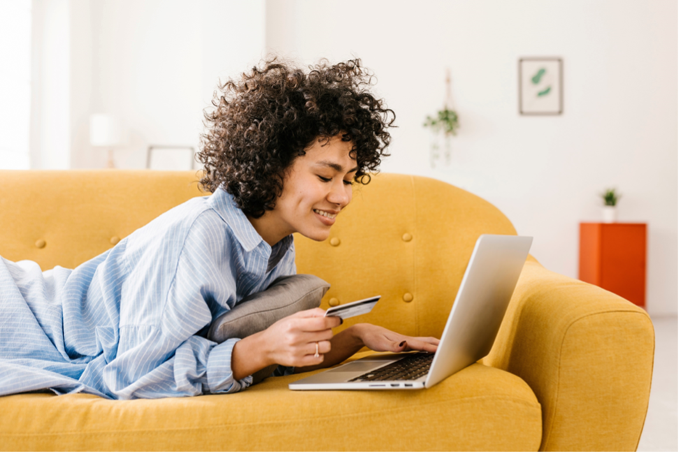 Una mujer con cabello rizado sonriendo, vestida de camisa Oxford azul, acostada en un sofá amarillo. Está trabajando en una computadora portátil encima del colchón del sofá y sostiene lo que parece ser una tarjeta de crédito en la mano derecha.