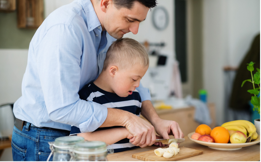 Una imagen de un niño joven con cabello rubio con un hombre con cabello marrón vestido de camisa azul. Los dos individuos están en la cocina, el adulto parado detrás del niño, ayudándolo a cortar un banano con un cuchillo guiándolo para asegurar que esté cortando el banano de manera segura.