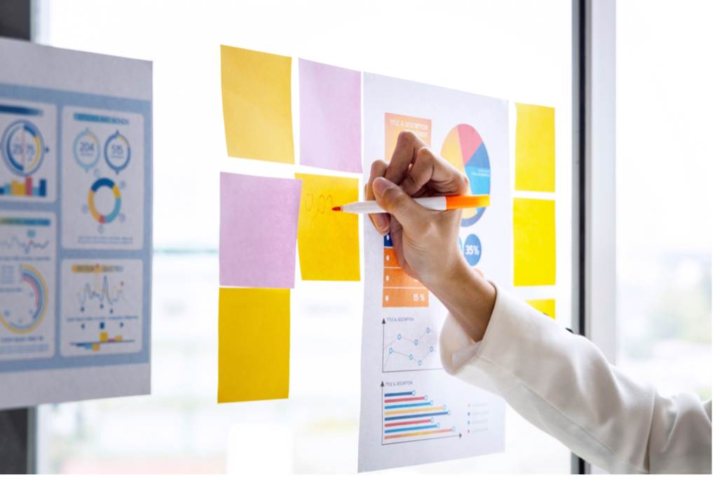 An image of pink and yellow sticky notes and paper with graphs on it stuck to a board. A person’s arm is in the image, and they are holding an orange marker writing something on one of the yellow sticky notes.