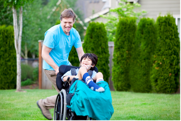 An image of a man in a blue shirt, smiling, while he pushes a young boy in a wheelchair. The boy has black hair and is wearing a blue shirt with dark blue socks. The young boy is laughing. The man and the boy are outside in a grassy area in the backyard of a home, surrounded by trees.