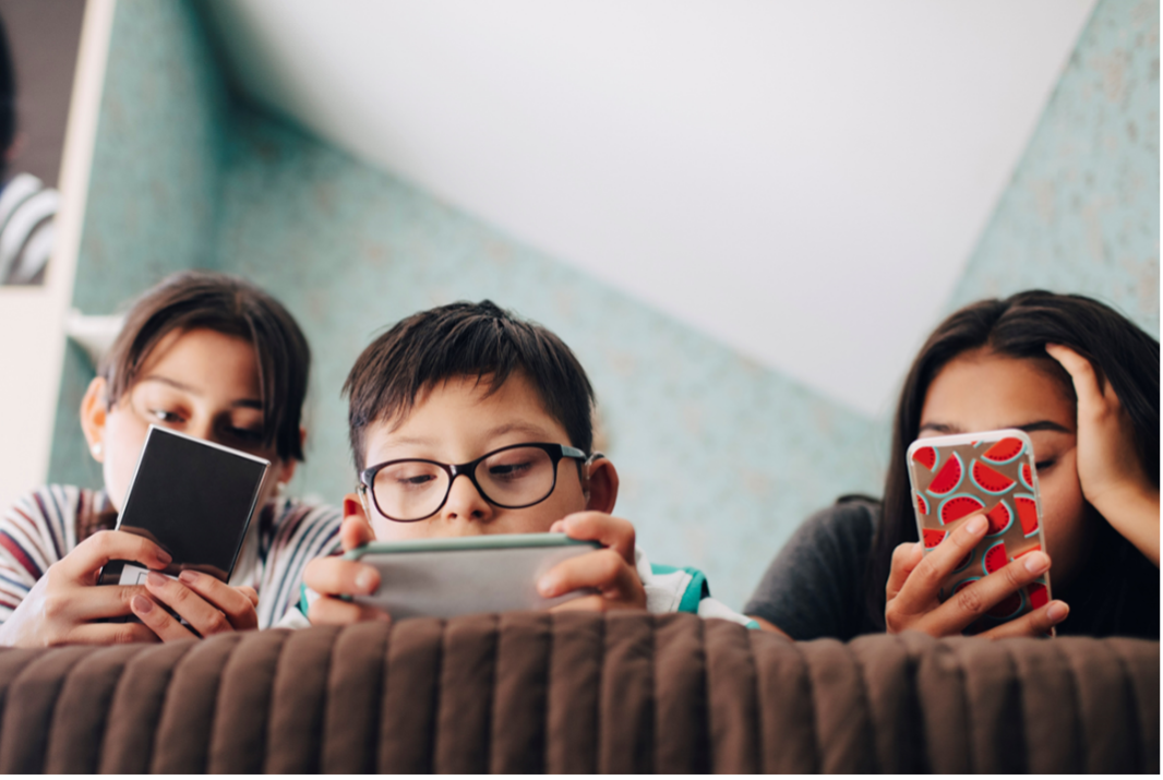 An image of three children, resting on a bed or couch, each holding a smart phone, looking at the screens. The image is zoomed in on their faces and the background is blurred.