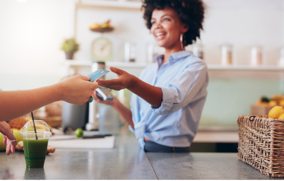 An image of a woman behind a service counter at a business, wearing a blue oxford shirt, taking a payment from an individual whose face is not pictured, giving her a credit card to pay for an item.
