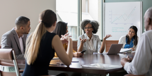 A group of professionals engaged in a discussion around a conference table, with a whiteboard displaying a line graph in the background.