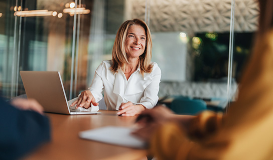 A woman sits at a table with a laptop. She smiles and engages in discussion with two people across from her in a bright office setting.